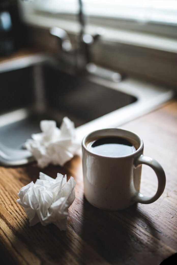 A close-up of a half-empty lukewarm coffee mug on a cluttered kitchen table, surrounded by crumpled tissues and crumbs, capturing a quiet moment of regret.