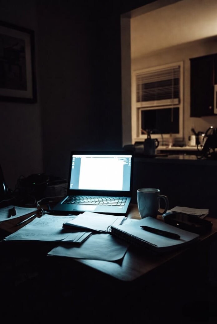 Chaotic night desk in an American kitchen, laptop glowing amid scattered notes and coffee.