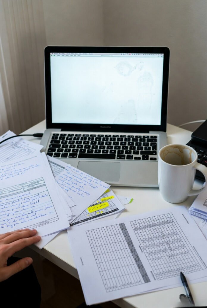 Messy desk from above, tilted phone shot: chaotic spreadsheet on glaring screen, cold coffee rings, scattered notes, stressed 2pm vibe.