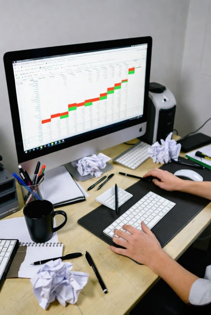 Chaotic desk mid-meltdown: frantic hands on keyboard, crumpled papers, endless spreadsheet refresh.
