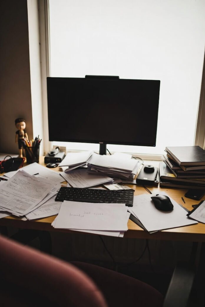 Low-angle shot of chaotic desk, keyboard buried under notes, empty screen glowing faintly