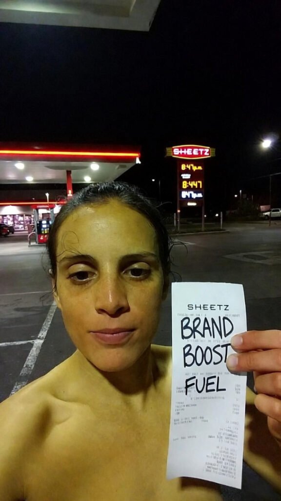Sodium-lit parking lot selfie, exhausted grin, hand raising receipt marked "BRAND BOOST FUEL" in Sharpie.