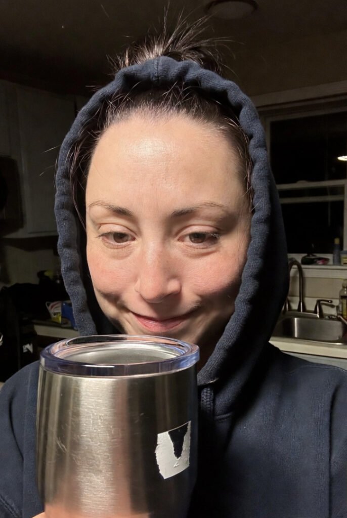 Flash-lit kitchen selfie: hoodie up, messy bun, holding sticker-peeling Yeti, slight smile