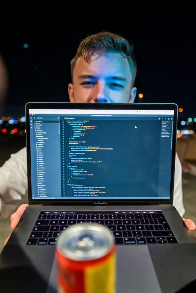 Selfie-style night shot: person squinting at glowing MacBook Pro, blue light on face, empty energy drink can in foreground.