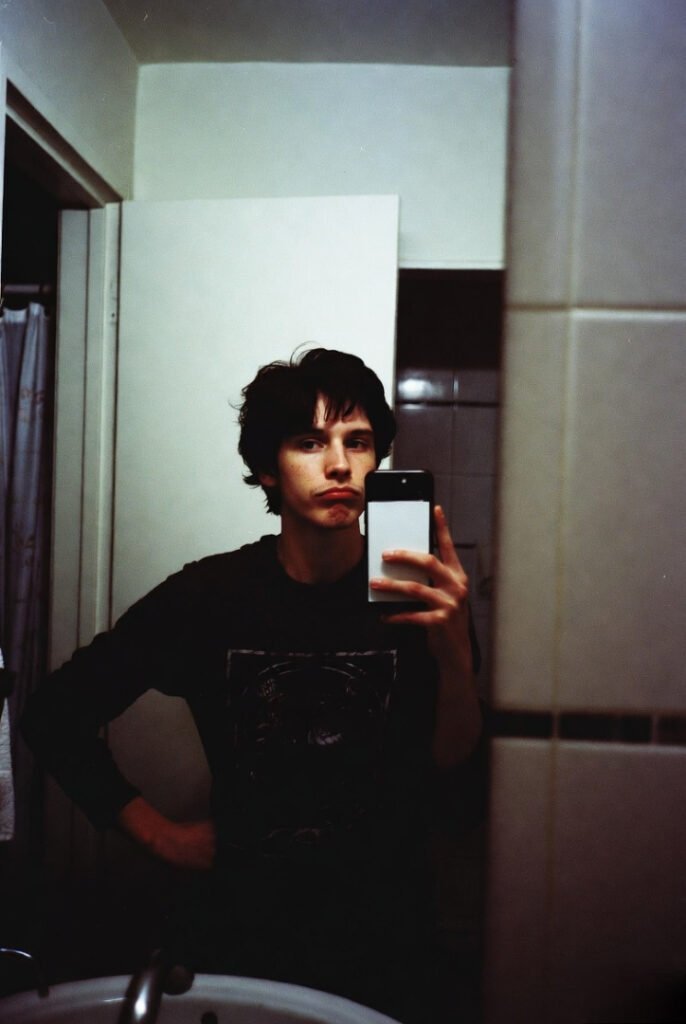 Grainy bathroom mirror selfie: messy hair, tired eyes, half-proud half-defeated expression after late-night cold pizza.